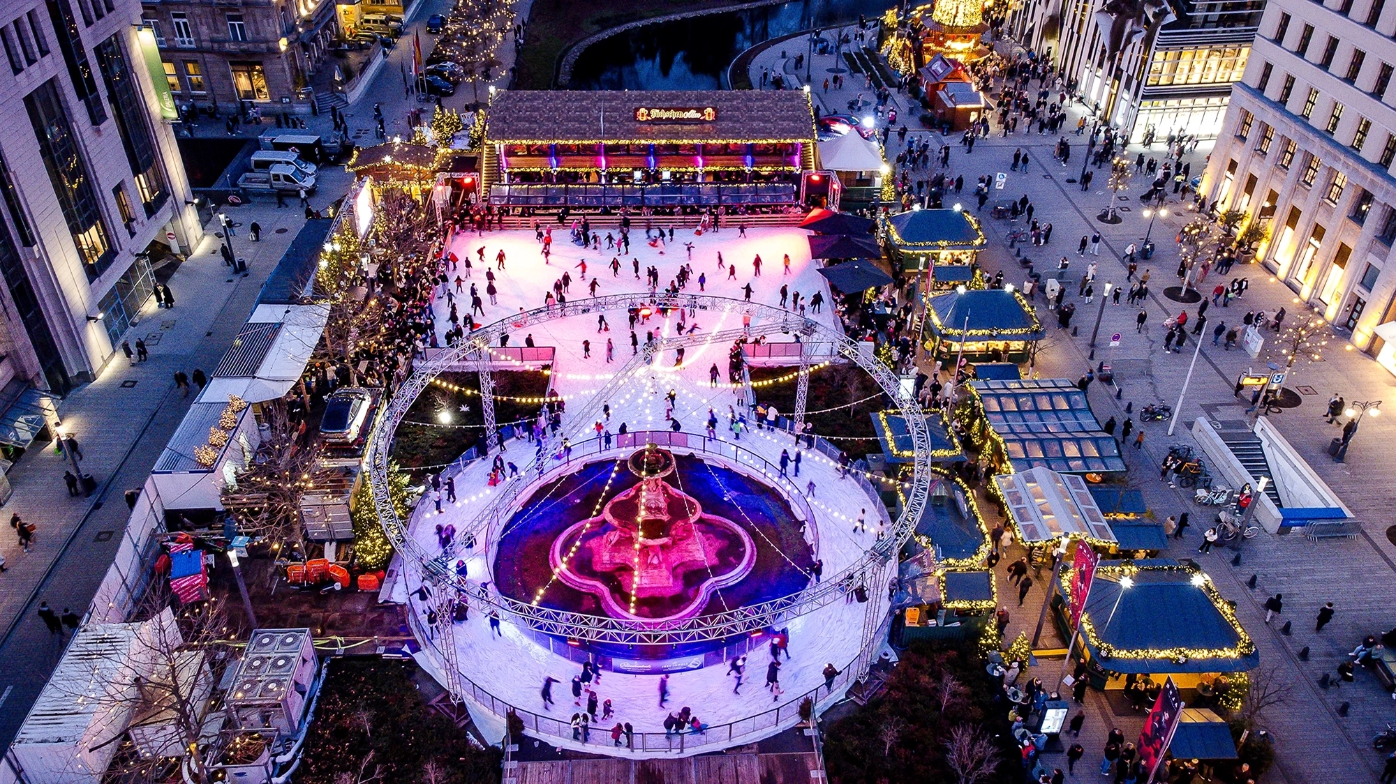 Drohnenfoto auf Koe-on-ice in Richtung Hofgarten mit Blick auf die zentrale Eisfläche, die Fuechschen-Alm, dem seitlichen Weihnachtsmarkt mit den klassischen grünen Pavillons und im Hintergrund der Köbogen